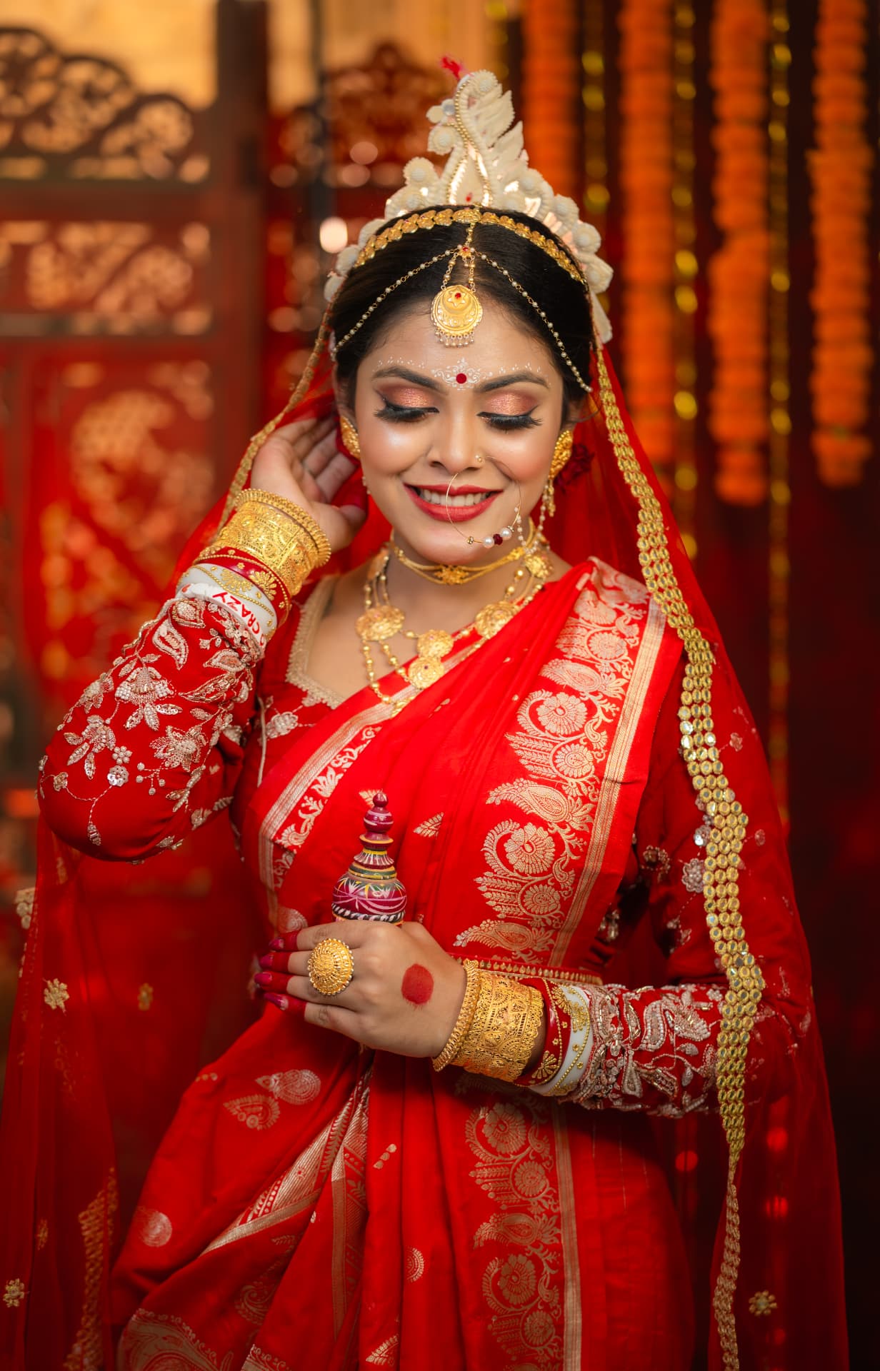 Smiling bride in a red benarasi with a white shola crown, gold jewellery and a warm marigold backdrop