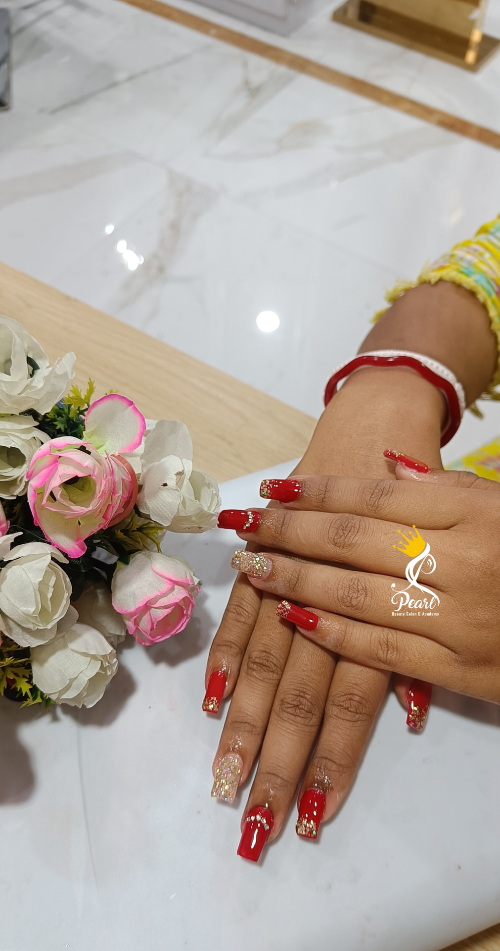Hands with glossy red square-tip nail extensions with gold glitter accents beside white and pink roses